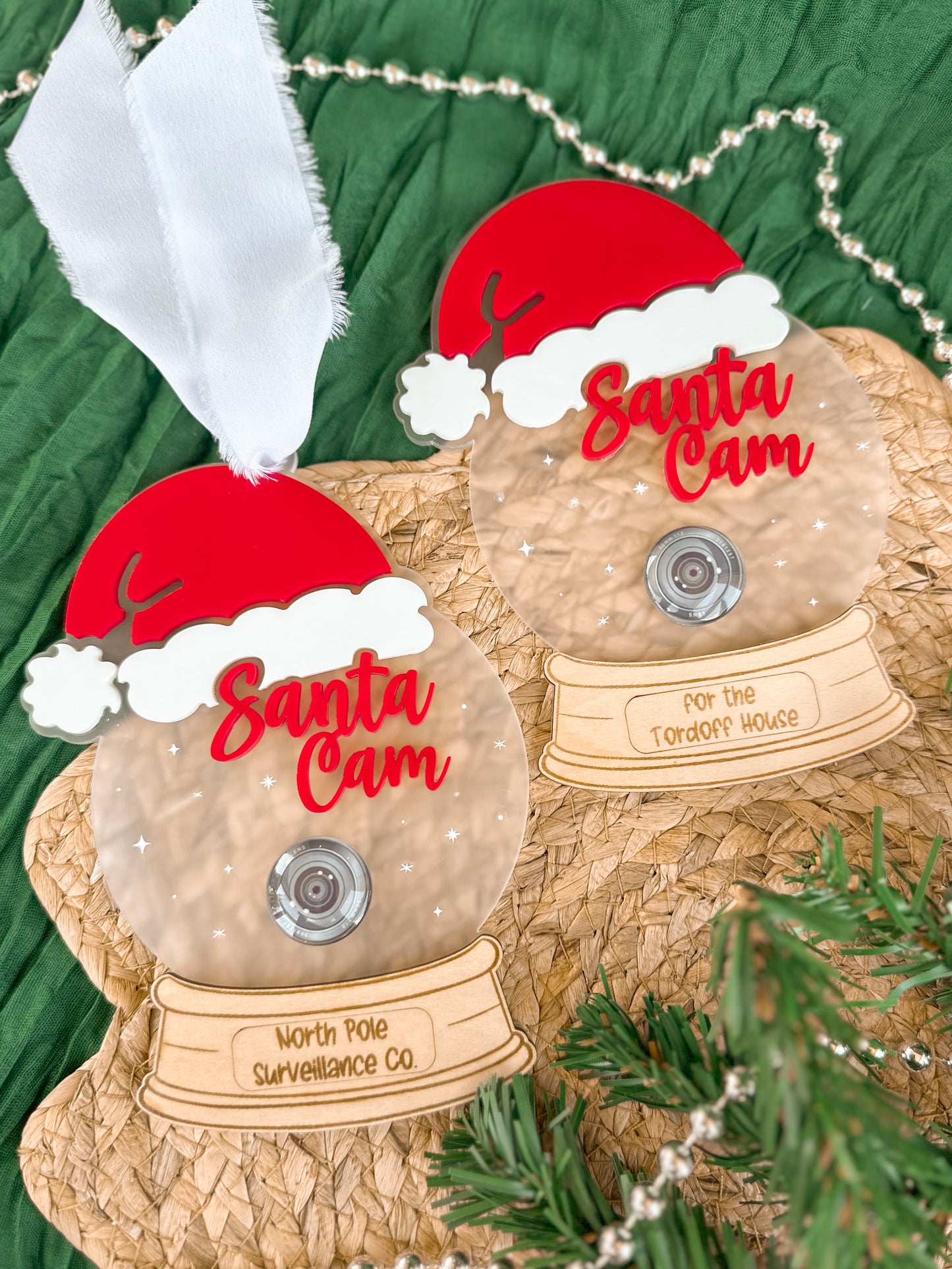 Two 'Santa Cam' personalised Christmas decorations. Decorative acrylic ornament resembling a 'Santa Cam' with a red hat and clear frosted acrylic base with a wooden engraved bottom. Featuring stars and a realistic glass camera lens.