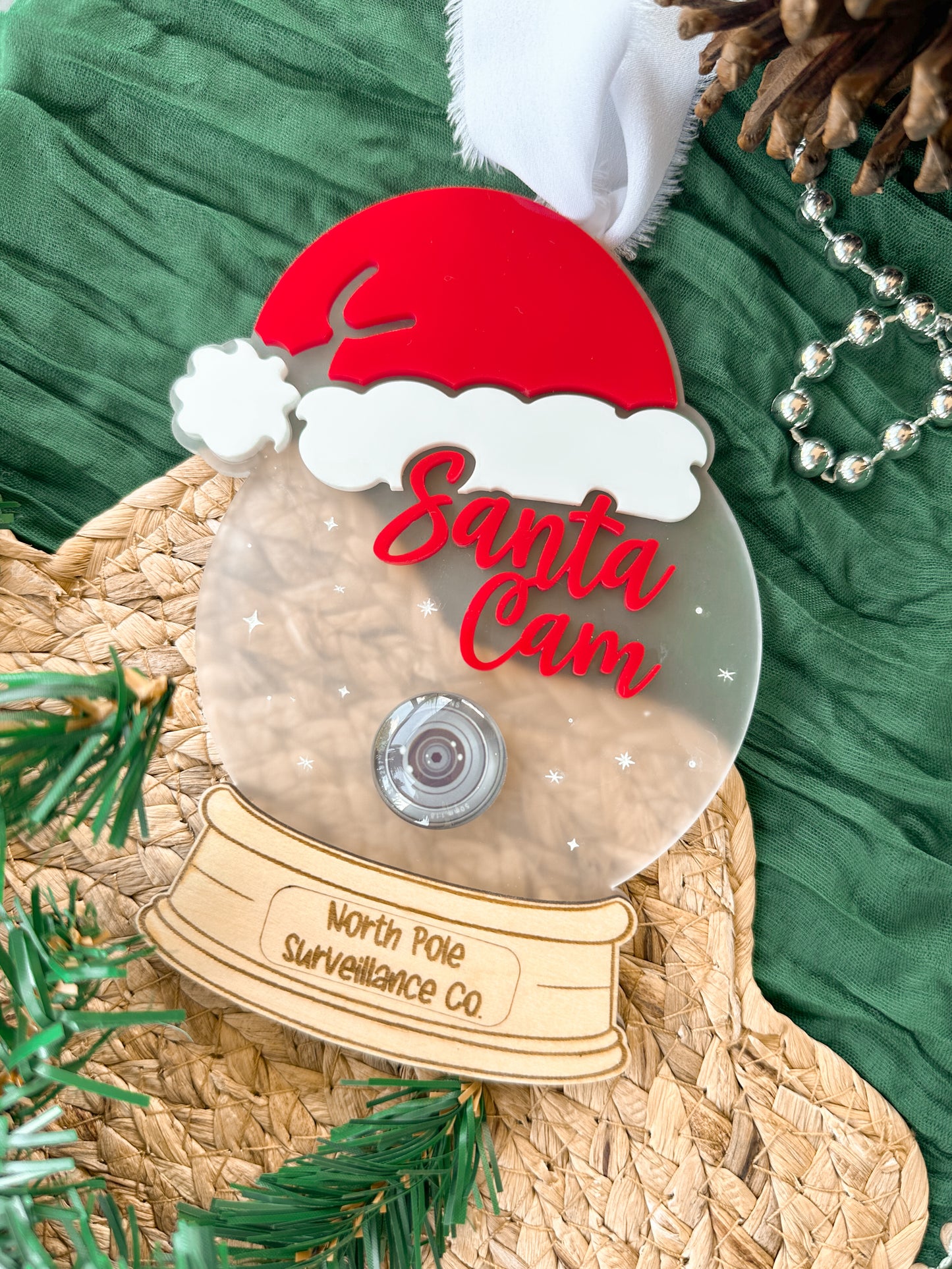 Decorative acrylic ornament resembling a 'Santa Cam' with a red hat and clear frosted acrylic base with a wooden engraved bottom. Featuring stars and a realistic glass camera lens.
