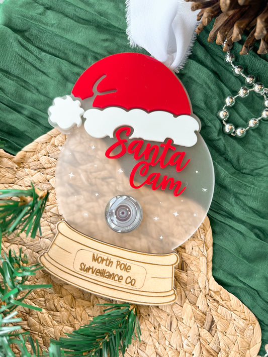 Decorative acrylic ornament resembling a 'Santa Cam' with a red hat and clear frosted acrylic base with a wooden engraved bottom. Featuring stars and a realistic glass camera lens.